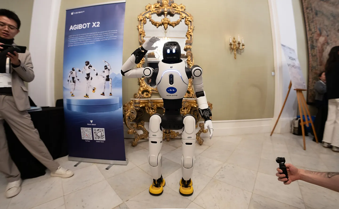 Den vita humanoiden AgiBot X2 står framför en mässbanderoll med texten AGIBOT X2.