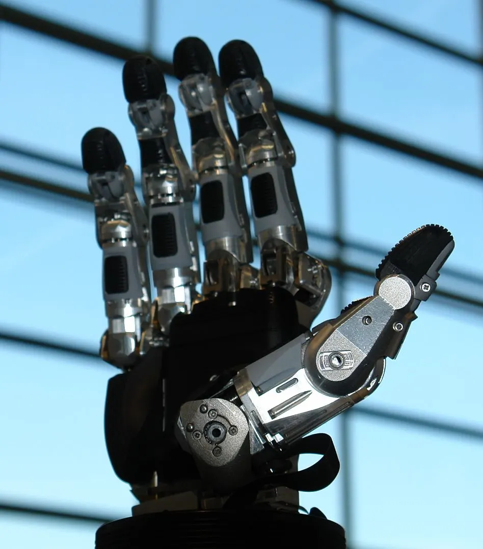 En femfingrad robothand i metall och plast med synliga leder och senor, mot ett fönster.