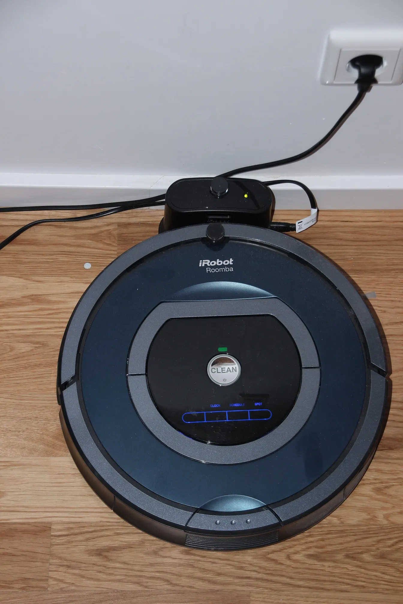 En mörk iRobot Roomba står i sin laddstation på ett trägolv.