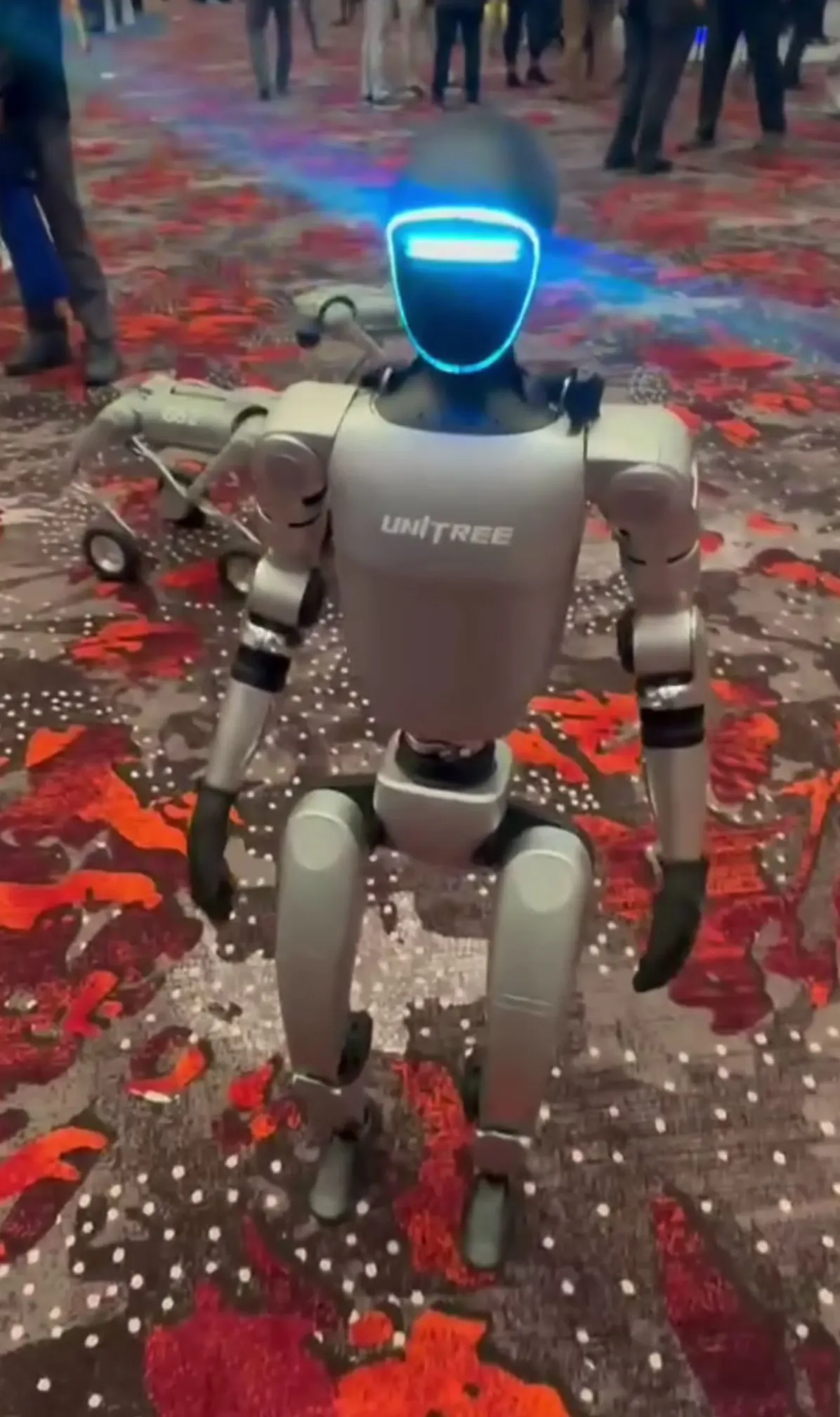 Den tvåbenta roboten Unitree G1 står på ett mönstrat golv på en mässa, med lysande blå ögonpanel.